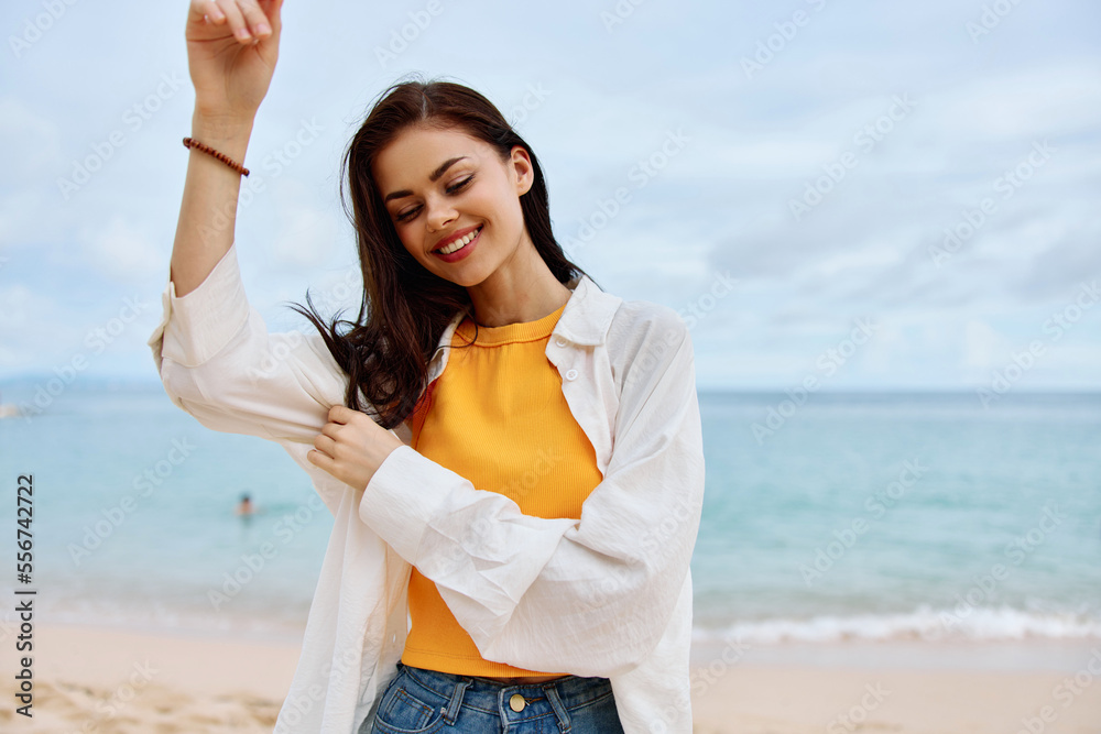 Happy woman smile with teeth with long hair brunette walks along the beach in a yellow t-shirt denim shorts and a white shirt near the sea summer journey and feeling of freedom, balance Happy woman smile with teeth with long hair brunette walks along the beach in a yellow t-shirt denim shorts and a white shirt near the sea summer journey and feeling of freedom, balance