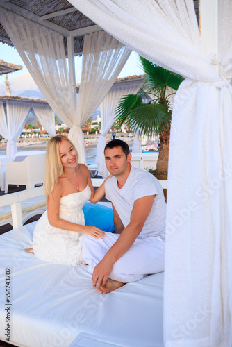 A young couple in a bungalow in the resort area.  A young blonde woman and a young dark-haired man on the seashore. People are dressed in white clothing.