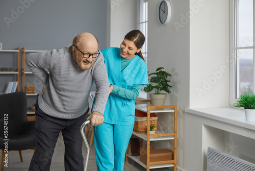 A young cheerful brunette nurse helping a senior patient to walk with his crutches. Physiotherapist helping old man in rehab. Health care worker helping an elderly man and smiling.