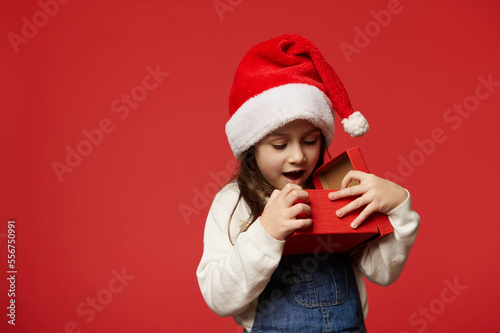 Delightful Caucasian 5 years old little child girl in Santa hat  expressing surprise and positive emotions while unpacking Christmas present over red background. Boxing Day. Happy New Year concept