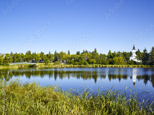 Tranquil Mirror Lake in the Camrose community with a footbridge from a park and the white Ukrainian Catholic church building reflected in the water; Camrose, Alberta, Canada