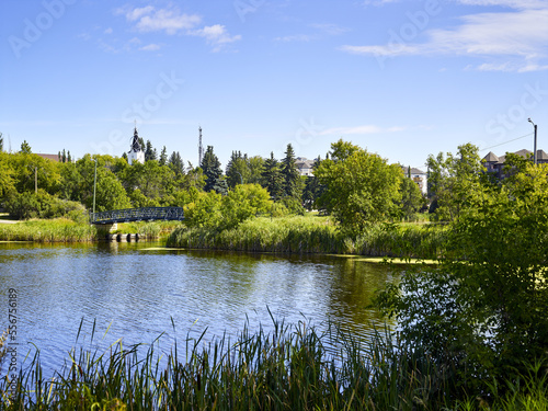 A tranquil Mirror Lake in the community of Camrose with a footbridge from a park and a white church tower seen through the treetops; Camrose, Alberta, Canada