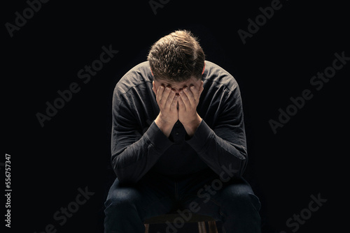 A young man sits with his face in his hands against a black background; Studio