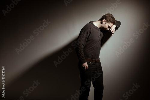 A young man stands leaning against a wall with his head on his arm against a studio background; Studio photo