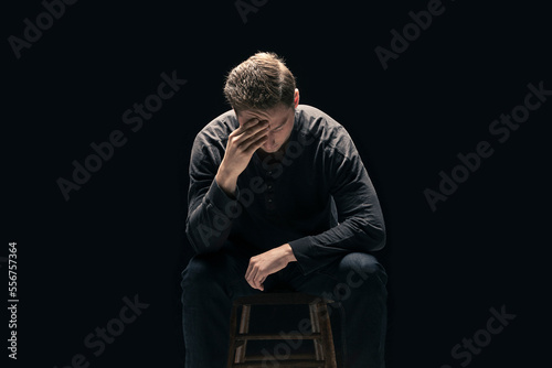 A young man sits with his face in his hands against a black background; Studio