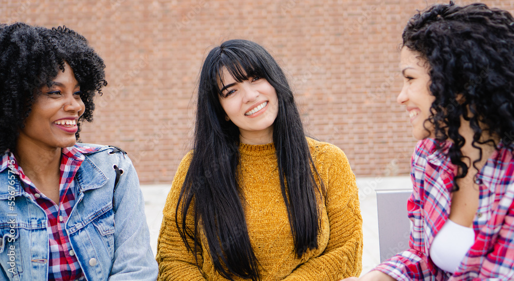 Fototapeta premium Three young, Latina and Hispanic interracial college friends chatting on a campus bench between classes.