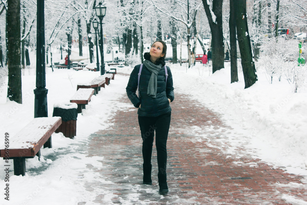 An attractive young brunette woman stands in snow-covered winter park smiles looking at a camera. Portrait of a beautiful girl. A path covered with white snow goes into distance. Cold weather in city.