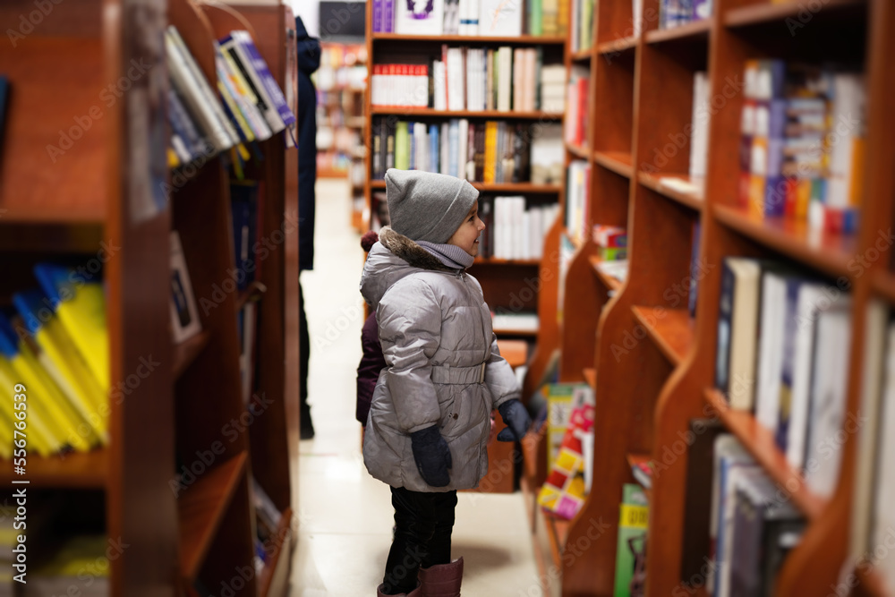 Little cute girl in jacket reaching a book from bookshelf at the library. Learning and education of european kid.