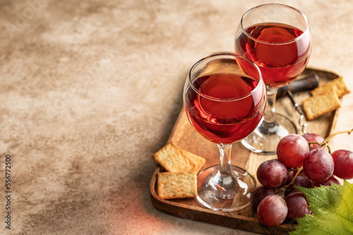 Two glasses of red wine, a bunch of red grapes, crackers and corkscrew on a wooden tray on beige textured background with text space. Party for two concept