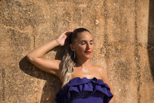 Portrait of beautiful young woman  with ponytail  wearing a purple dress  with her eyes closed and relaxed leaning against a stone wall receiving the sun rays. Concept beauty  fashion  relax  sunny.