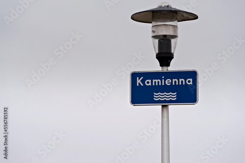Niebieska tabliczka z nazwą rzeki ( Kamienna) i piktogramem płynącej wody, zawieszona na latarni ulicznej ,na szarym tle nieba .