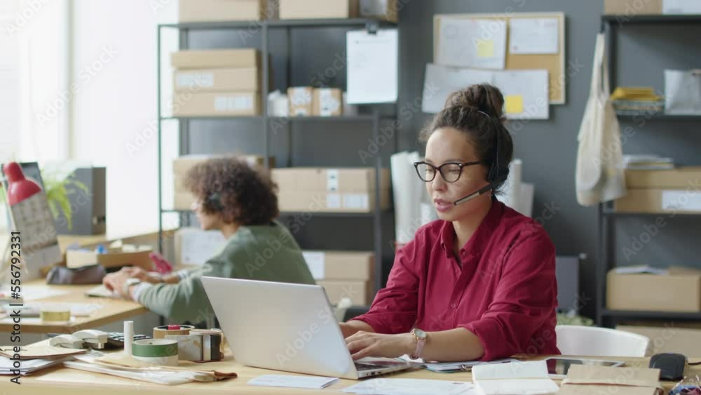 Female customer support agent in headset using laptop and speaking on web call while working in delivery service office Female customer support agent in headset using laptop and speaking on web call while working in delivery service office