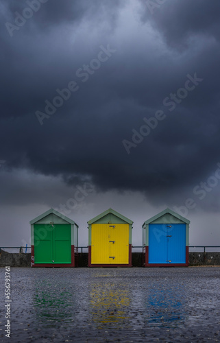 Three brightly colored beach huts sit beneath a storm cloud on a wet autumnal day; Hove, East Sussex, England, United Kingdom photo