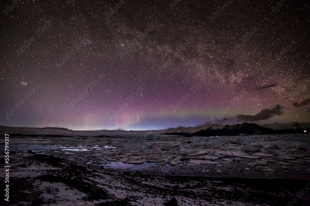 Aurora boreale all'orizzonte sormontata dalla via lattea da sinistra a destra con la laguna ghiacciata in primo piano e le montagne sullo sfondo