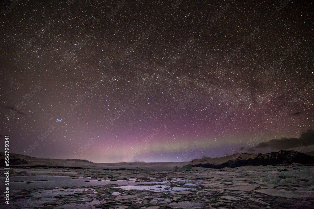 Aurora boreale all'orizzonte sormontata dalla via lattea con la laguna ghiacciata in primo piano e le montagne sullo sfondo