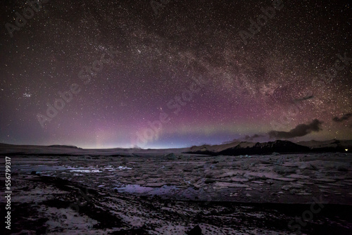 Aurora boreale all'orizzonte sormontata dalla via lattea da sinistra a destra con la laguna ghiacciata in primo piano e le montagne sullo sfondo photo