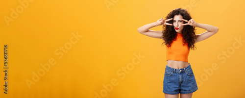 Portrait of good-looking daring and sexy stylish woman with curly hairstyle in red lipstick and cropped top showing victory or peace gestures overs eyes squinting folding lips flirty over orange wall