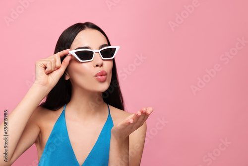 Beautiful young woman in stylish sunglasses blowing kiss on pink background. Space for text