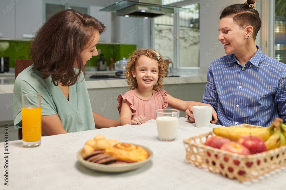 Happy little girl and her two mothers sitting at breakfast table