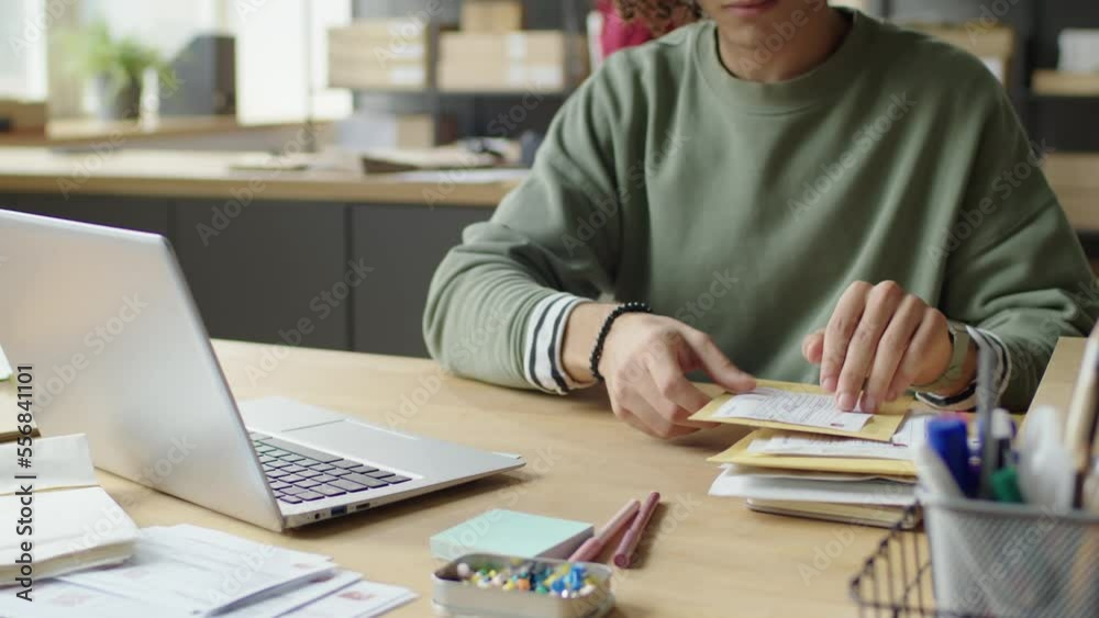 Tilt up shot of young male worker reading information on envelopes and using laptop while sorting mail in delivery service office
