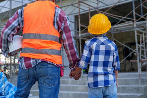 Male contractor holding boy's hand standing looking at building under construction, father taking son to look at his construction site, low view from behind, father and son holding hands.