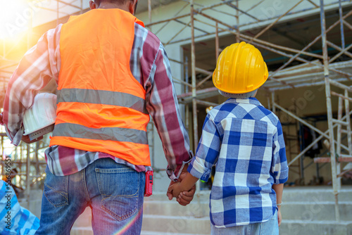 Male contractor holding boy's hand standing looking at building under construction, father taking son to look at his construction site, low view from behind, father and son holding hands.