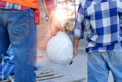 Male contractor holding boy\'s hand standing looking at building under construction, father taking son to look at his construction site, low view from behind, father and son holding hands.