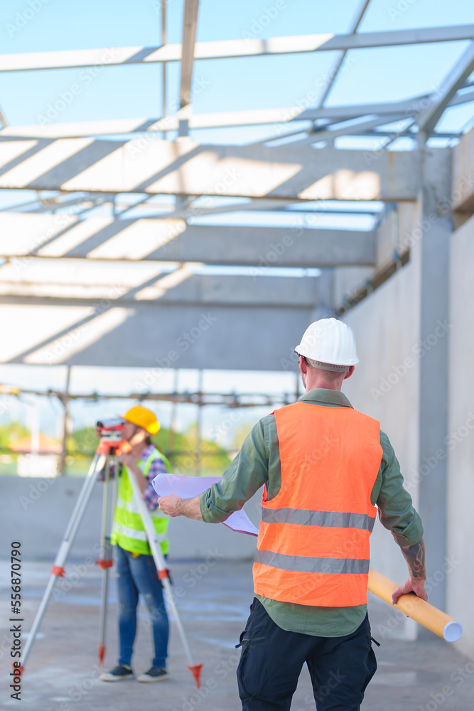 Construction Worker Using Theodolite Surveying Optical Instrument for Measuring Angles in Horizontal and Vertical Planes on Construction Site. Engineer and Architect Using blueprint Next to Surveyor. Construction Worker Using Theodolite Surveying Optical Instrument for Measuring Angles in Horizontal and Vertical Planes on Construction Site. Engineer and Architect Using blueprint Next to Surveyor.