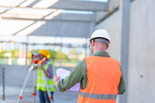 Construction Worker Using Theodolite Surveying Optical Instrument for Measuring Angles in Horizontal and Vertical Planes on Construction Site. Engineer and Architect Using blueprint Next to Surveyor.