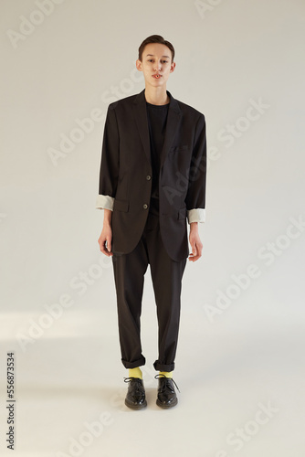 Pretty gender neutral model in stylish male suit looking at camera and blowing bubble from chewing gum while standing against gray background photo