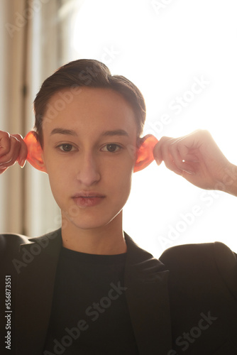 Attractive gender neutral model in suit looking at camera and pulling ears while standing against window on sunny day photo