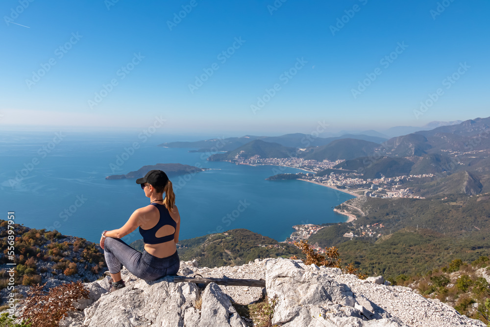Naklejka premium Hiking woman sitting on rock enjoying panoramic aerial view on coastline of Budva and Sveti Nikola Island seen from Goli Vrh, Adriatic Mediterranean Sea, Montenegro, Balkan, Europe. Budvanian Riviera