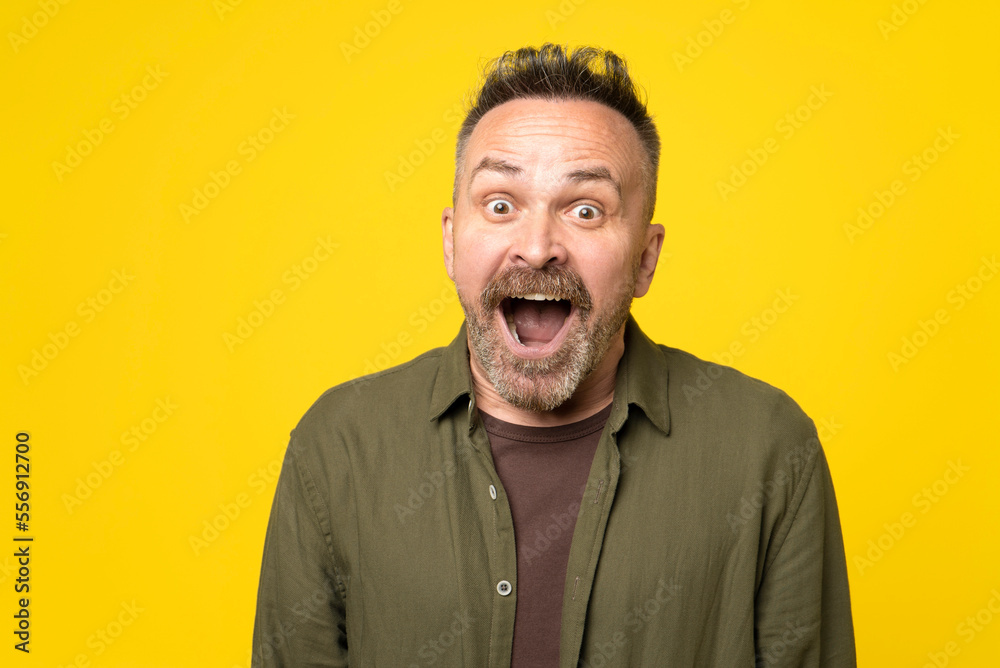 Excited bristle and mustached man 50s exclaims in happiness, recieved pleasant surprise, can`t believe his eyes, expresses great surprisment, isolated over yellow background