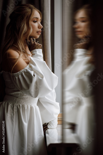 A stylish and pretty young Caucasian girl is standing by the window, looking away, side view. lush hairstyle and make-up. wears a white dress