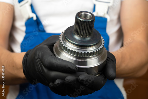 Auto mechanic in blue jumpsuit holds in his hands hinge of equal angular velocities of car and rotates it in black gloves. . Car wheel drive replacement concept, auto parts.