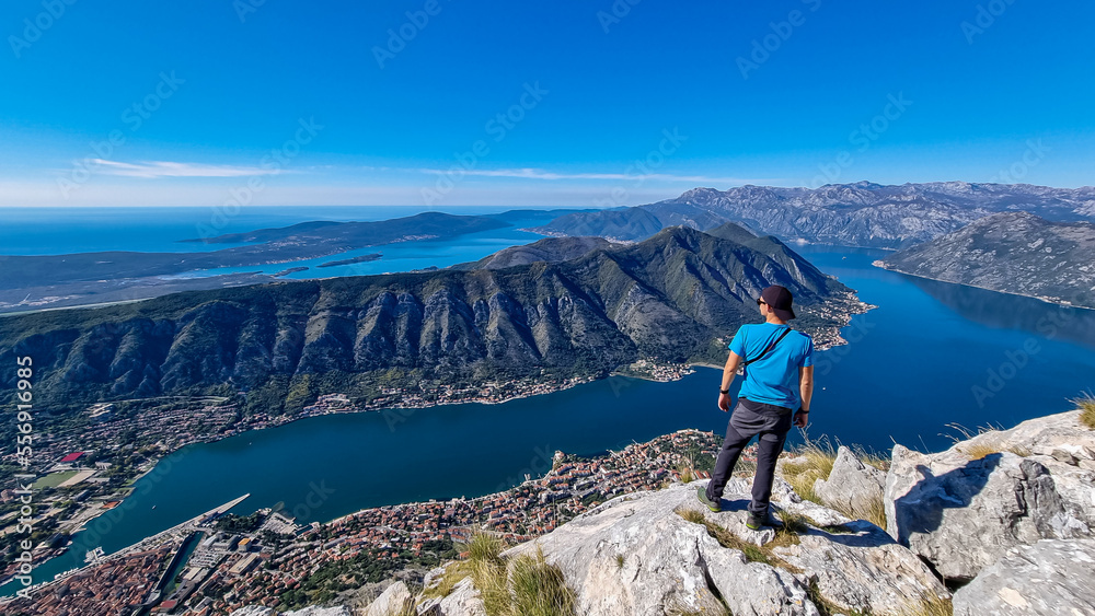 Man with panoramic view from Pestingrad (Derinski Vrh) of Kotor bay in sunny summer, Adriatic Mediterranean Sea, Montenegro, Balkan, Europe. Fjord winding along coastal towns. Lovcen, Orjen mountains