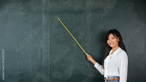 Back to school concept. Happy beautiful young woman standing hold pointer to back board, Asian female teacher smiling with wooden stick pointing to blackboard at school in classroom, Education