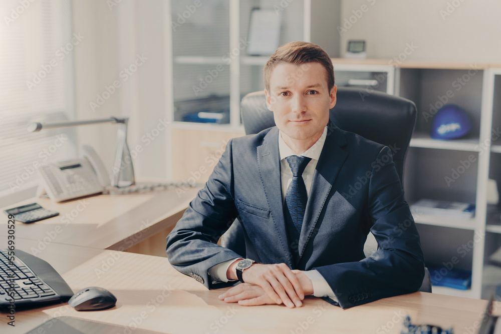 Indoor shot of pleasant looking male manager in black suit, sits in office, has confident expression at camera, works during working day, makes decision concerning future plans. CEO in cabinet Indoor shot of pleasant looking male manager in black suit, sits in office, has confident expression at camera, works during working day, makes decision concerning future plans. CEO in cabinet
