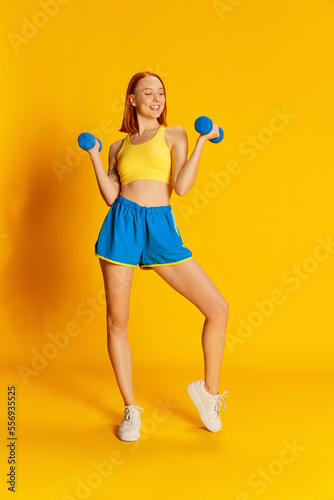 Portrait of young redhead girl in sportswear training with sports equipment over yellow background. Hands exercises