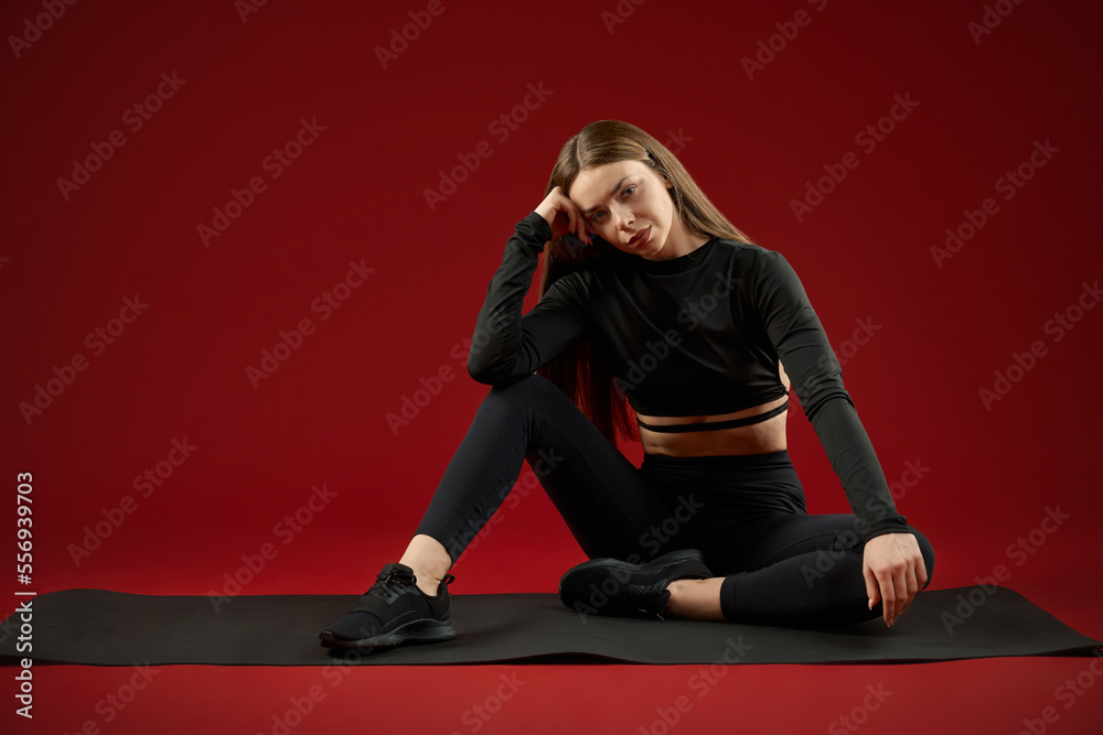 Beautiful seriously brunette woman wearing in black sport suite sitting resting after training on идфсл yoga mat. Sporty fit female lean on knee looking at camera, relaxing on floor on red background.