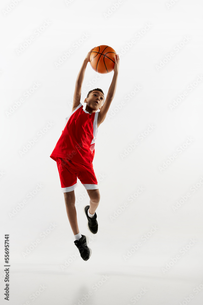 Portrait of boy in red uniform training, playing basketball in a jump over grey studio background