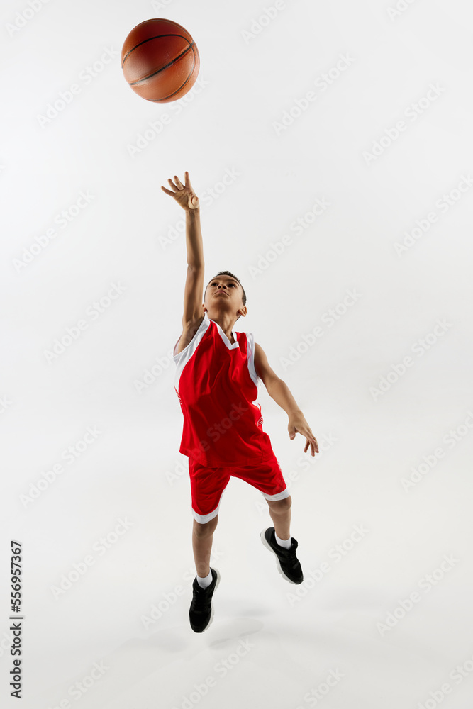 Portrait of boy in red uniform training, playing basketball over grey studio background. Throwing ball