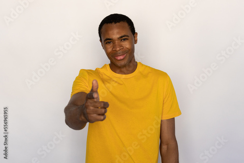 Portrait of young man pointing at camera and sneering against white background. African American guy wearing yellow T-shirt looking at camera and smiling. Mockery concept
