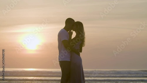 Romantic Caucasian couple kissing against sunset background while standing at seashore. Silhouettes of athletic man and long-haired pretty lady hugging, sharing happy moments together. Romance concept