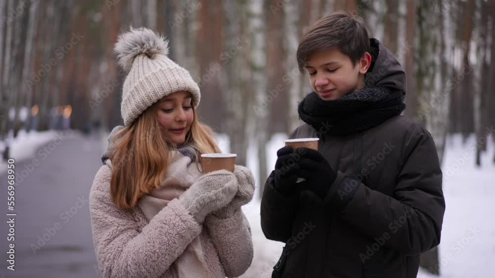 Smiling relaxed teen couple standing with hot tea cups on park alley talking. Portrait of carefree positive Caucasian teenage boy and girl chatting enjoying winter weekend outdoors. Slow motion Smiling relaxed teen couple standing with hot tea cups on park alley talking. Portrait of carefree positive Caucasian teenage boy and girl chatting enjoying winter weekend outdoors. Slow motion