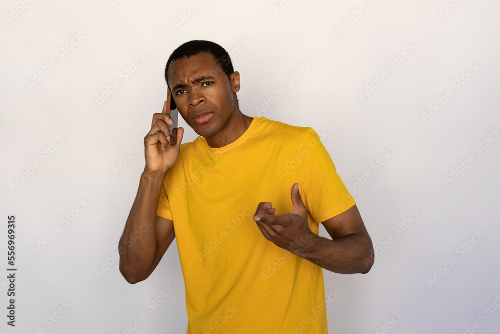 Portrait of frowning young man talking on smartphone over white background. African American guy wearing yellow T-shirt holding mobile phone and looking at camera. Mobile communication concept Portrait of frowning young man talking on smartphone over white background. African American guy wearing yellow T-shirt holding mobile phone and looking at camera. Mobile communication concept