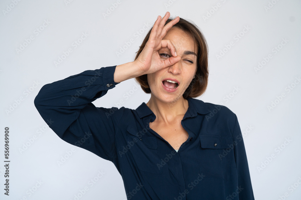 Surprised woman looking through okay gesture. Female model in dark blue shirt making ok gesture with open mouth. Portrait, studio shot, amazement concept Surprised woman looking through okay gesture. Female model in dark blue shirt making ok gesture with open mouth. Portrait, studio shot, amazement concept