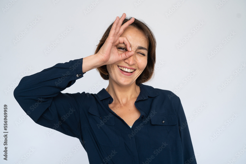 Cheerful woman looking through okay gesture. Female model in dark blue shirt making okay gesture and smiling. Portrait, studio shot, fun concept Cheerful woman looking through okay gesture. Female model in dark blue shirt making okay gesture and smiling. Portrait, studio shot, fun concept