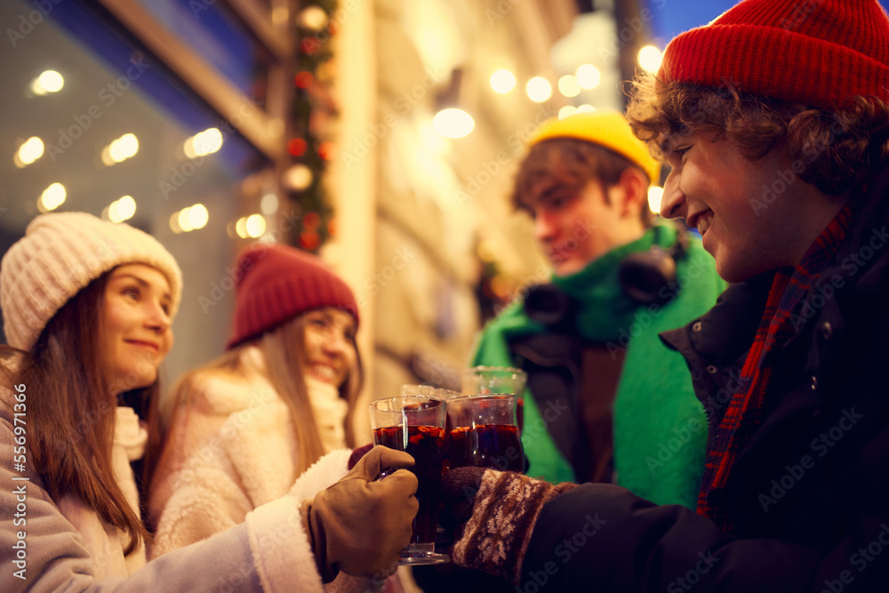 Group of young people, boys and girls spending time together at festive Christmas street, tasting hot mulled wine and coffee. Concept of winter holidays, emotions, friendship Group of young people, boys and girls spending time together at festive Christmas street, tasting hot mulled wine and coffee. Concept of winter holidays, emotions, friendship