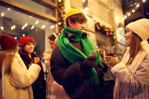 Happy young men and girls, friends walking at festive Christmas street and drinking mulled wine. Concept of holidays, winter, New Years fairs, friendship